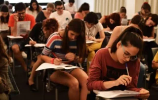 Un grup nombrós d'estudiants asseguts en files en una aula, concentrats fent un examen. Cadascun disposa d'un petit escriptori acoblat a la cadira i està utilitzant paper i bolígraf.