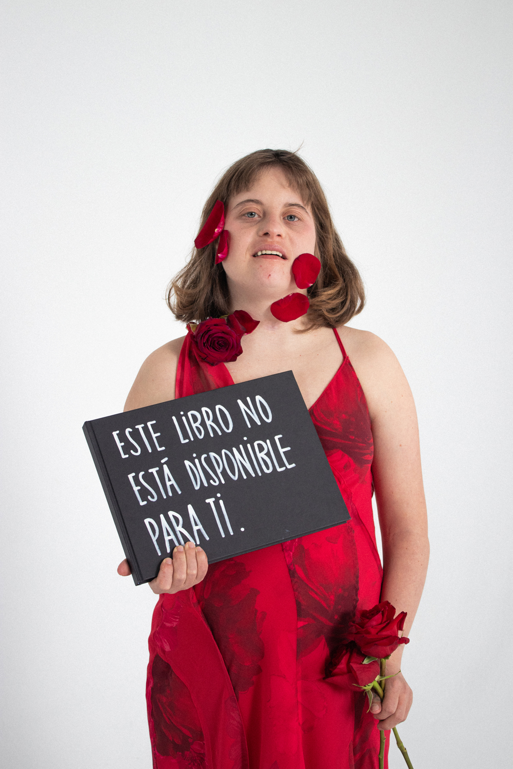 Clara-Teatre Sense Límits-EÒLIA CSAD Una dona vestida de vermell amb pètals de rosa a la cara sosté un cartell que diu "Aquest llibre no està disponible per a tu" sobre un fons blanc.