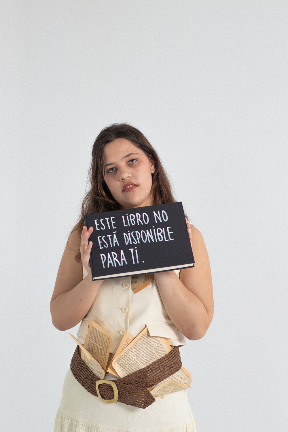 Martina-Teatre-Sense-Límits-EÒLIA-CSAD Una dona de cabell castany i rostre seriós subjecta un llibre negre amb text en castellà que diu: "ESTE LIBRO NO ESTÁ DISPONIBLE PARA TI.". Porta un armilla de color crema i un cinturó de corda amb papers vells que hi pengen.
