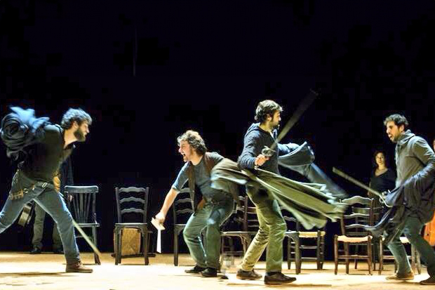 Rei Lear Teatre Lliure Una escena de lluita a l'escenari amb quatre homes. Dues figures a l'esquerra s'enfronten a dues de la dreta, totes armades amb pals, banyes o teles onejant. Al fons hi ha diverses cadires negres de fusta.