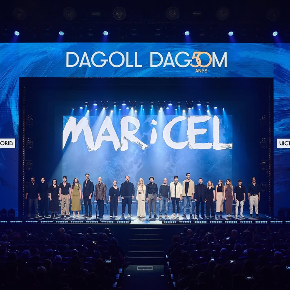 Fotografia d'un escenari de teatre buit amb un gran fons de pantalla amb un disseny abstracte de colors blaus marins i el text "DAGOLL DAGOM 50 ANYS" al capdamunt. Al centre de l'escenari, una gran pantalla mostra el títol del musical "MAR I CEL" en lletres grans i blanques. A sota, tot l'elenc i l'equip de l'última producció de Mar i Cel estan drets en una línia àmplia. Són aproximadament 25-30 persones, majoritàriament vestides de negre o colors neutres, mirant cap al públic o cap endavant.