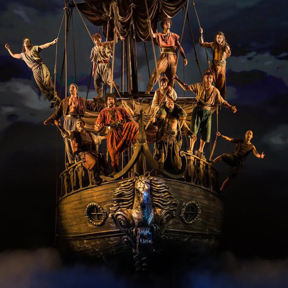 Una foto d'escena gran angular i molt espectacular que captura la majestuositat del vaixell pirata del musical Mar i Cel de Dagoll Dagom. El vaixell, amb una imponent proa tallada que inclou una figura de lleó o griu i un disseny molt detallat, es presenta en una posició elevada. Un gran nombre d'actors, vestits com a pirates amb robes marrons, ocres, vermelles i blaves d'època, estan dispersos per tota l'estructura.