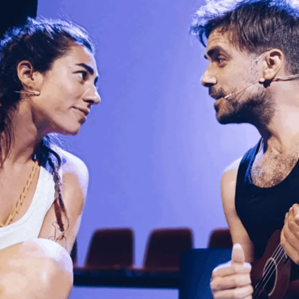 Foto d'escena que captura un moment íntim i d'interacció entre dos personatges de l'obra de teatre musical De amor nadie muere. A la dreta, Pol Forment amb barba, armilla de tirants negra i un micròfon de diadema, sosté un petit ukelele. Mira intensament la dona que té al costat amb una expressió concentrada i comunicativa.