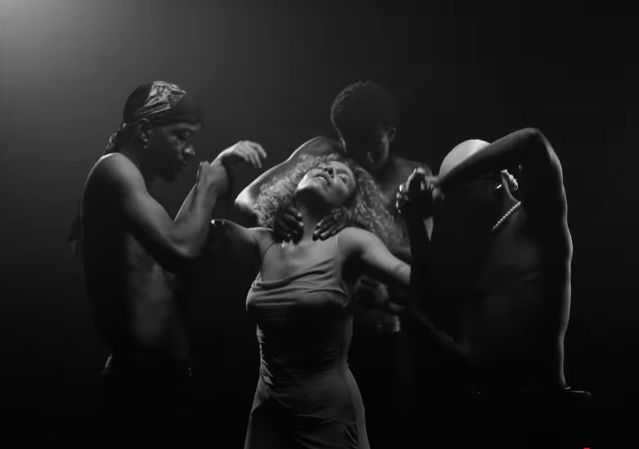 Fotografia en blanc i negre d'alt contrast amb il·luminació dramàtica que mostra una dona al centre, vestida amb un slip dress i amb el cap inclinat cap enrere, en una posició d'èxtasi o alliberament. Tres homes amb el tors nu l'envolten, tocant-la i formant un grup compacte. L'matge, molt fosca amb focus de llum intensa a les figures.