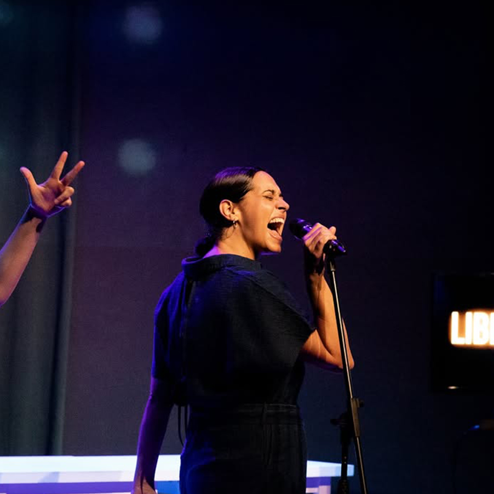 Júlia Cortina, canta amb el micròfon a prop de la boca en una intensa expressió facial. La figura es troba lleugerament girada cap a l'esquerra, amb la boca oberta i els ulls tancats o gairebé tancats, transmetent un gran esforç o passió vocal. Vesteix una granota o un vestit fosc de màniga curta.
