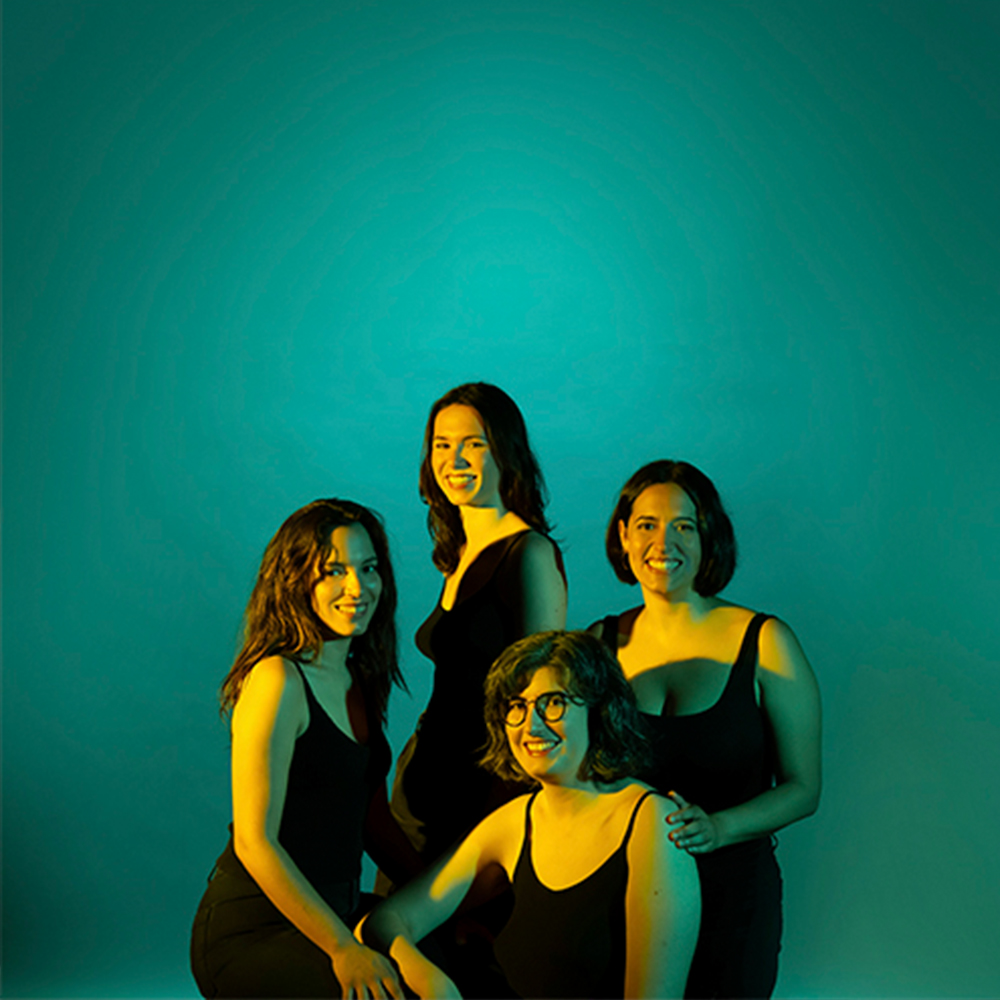 Retrat de quatre dones sobre un fons vibrant de color blau-verd (cian) il·luminat amb un llum de color groc que crea un fort contrast i un efecte de colorit dramàtic. Les quatre figures estan vestides de negre, amb tirants fins o màniga curta, suggerint un vestuari minimalista. Les dones es troben en un arranjament tancat i proper: dues estan dempeus a la part posterior, i dues estan assegudes o ajupides al primer pla. Totes miren directament a la càmera amb expressions serenes i somrients, transmetent confiança i camaraderia.