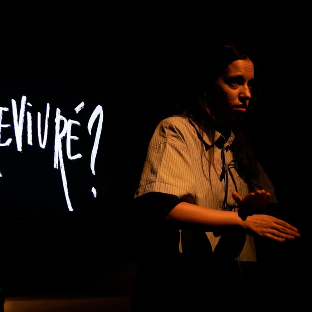 L'actriu Júlia Cortina, es troba al centre-dreta d'un escenari amb un fons completament fosc. Està il·luminada per una llum lateral o superior que enfosqueix parcialment la seva cara, però que il·lumina les seves mans en primer pla. Vesteix una camisa de ratlles verticals clares i fosques.