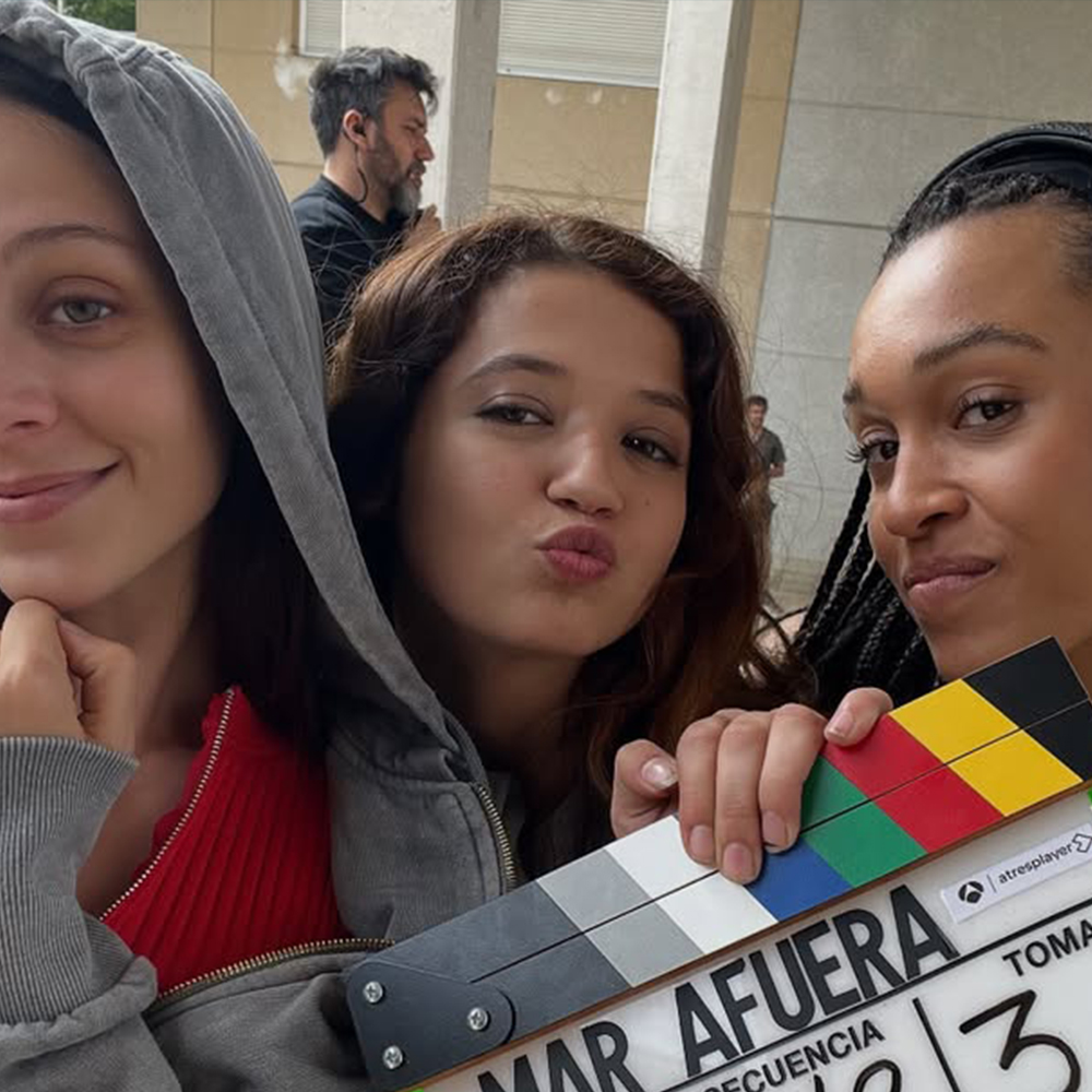 Tres dones durant una pausa o entre presa i presa en un set de rodatge exterior. A l'esquerra, una dona amb dessuadora gris i samarreta vermella somriu lleugerament. Al centre, una dona amb el cabell castany i una dessuadora gris posa fent un petó ("duck face"). A la dreta, una dona amb trenes i una expressió seriosa també mira a càmera.