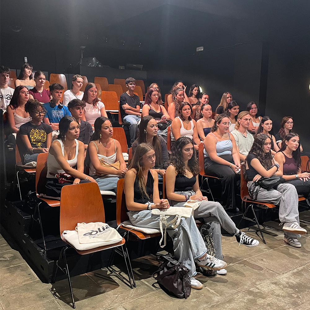 Una fotografia presa des de la part frontal i lleugerament elevada d'un teatre, mostrant un auditori amb un públic jove assegut en seients taronges de fusta i metall, organitzats en fileres ascendents. La sala és fosca, i la llum principal prové de la part superior del públic.