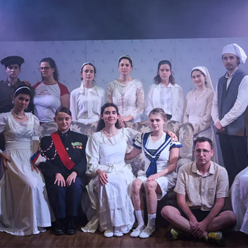 Fotografia d'un gran repartiment teatral de tretze persones posant formalment, com en un retrat de família d'època. Els intèrprets, majoritàriament dones, vesteixen amb una barreja de roba clàssica i elements més excèntrics, com ara un uniforme militar i un vestit de mariner. Es situen en tres nivells, utilitzant un sofà d'estil antic com a accessori.