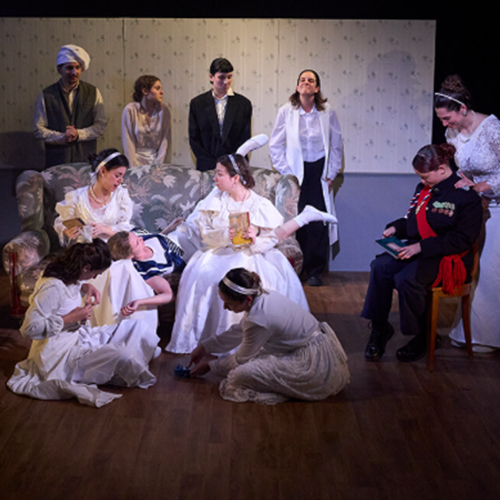Una escena teatral amb un gran repartiment d'onze intèrprets en un escenari interior amb un fons de paper pintat antic de color gris clar i disseny floral subtil. Tota la companyia està vestida amb un vestuari d'època que evoca el segle XIX o principis del XX. Hi ha un sofà d'estil vintage darrere, i els actors es troben en diverses posicions: alguns asseguts, altres drets i alguns ajaguts o interactuant a terra.