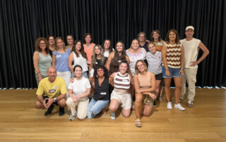 Foto grupal d'unes vint persones, la majoria dones, a sobre d'un escenari amb un terra de fusta clara i un gran teló de fons negre arrugat. El grup està dempeus i ajupit, formant dues files. Les persones de la fila del darrere estan dempeus, i les de la fila del davant estan ajupides.