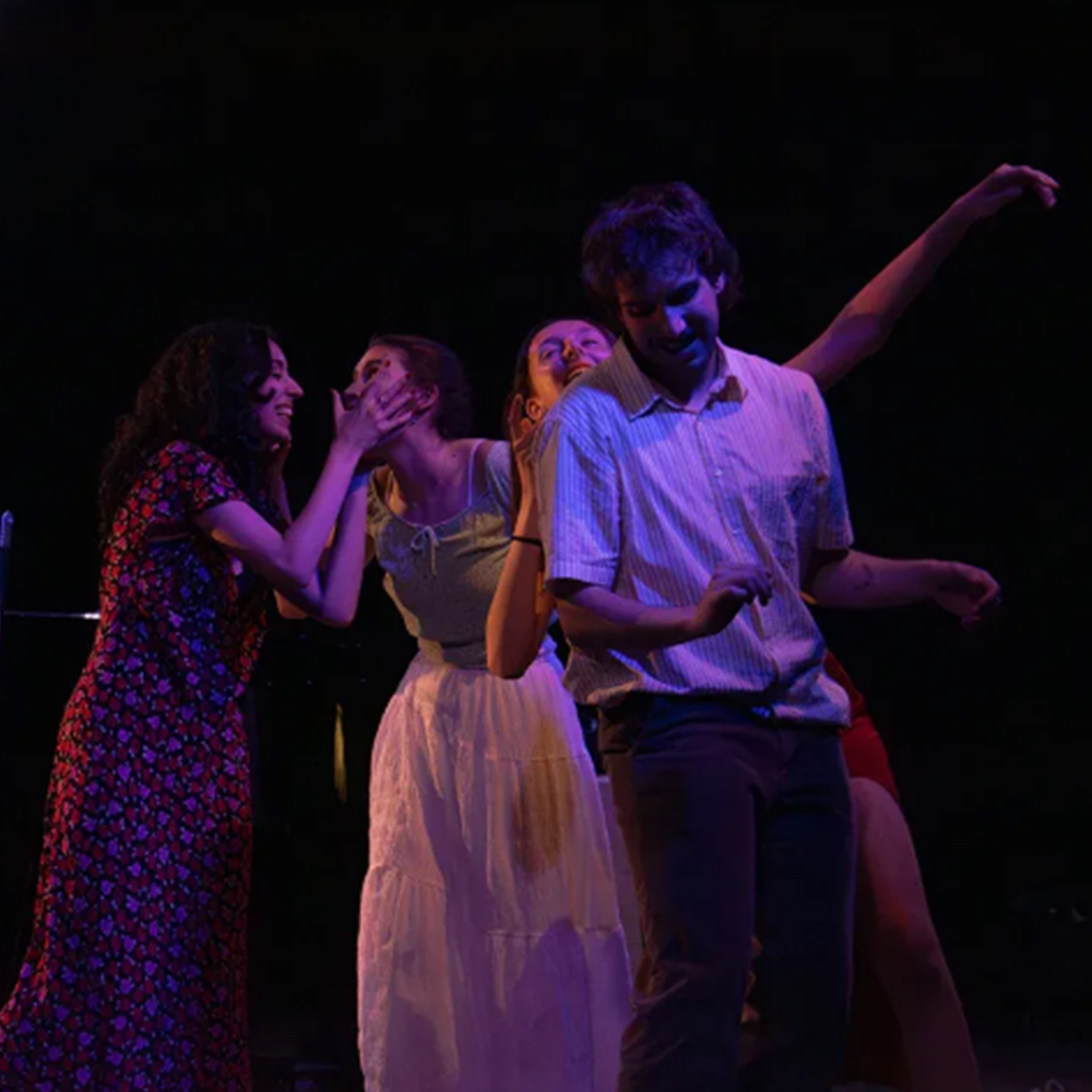 Quatre intèrprets en un escenari fosc, il·luminats amb una llum càlida i dramàtica que ressalta els seus rostres i expressions intenses. Dues dones a l'esquerra interactuen amb gests afectuosos i expressius: la dona de l'extrem esquerre, amb un vestit llarg floral vermell i negre, subjecta la cara de l'altra, que porta un vestit blanc o color crema d'estil vintage, i ambdues riuen o expressen un gran impacte emocional.