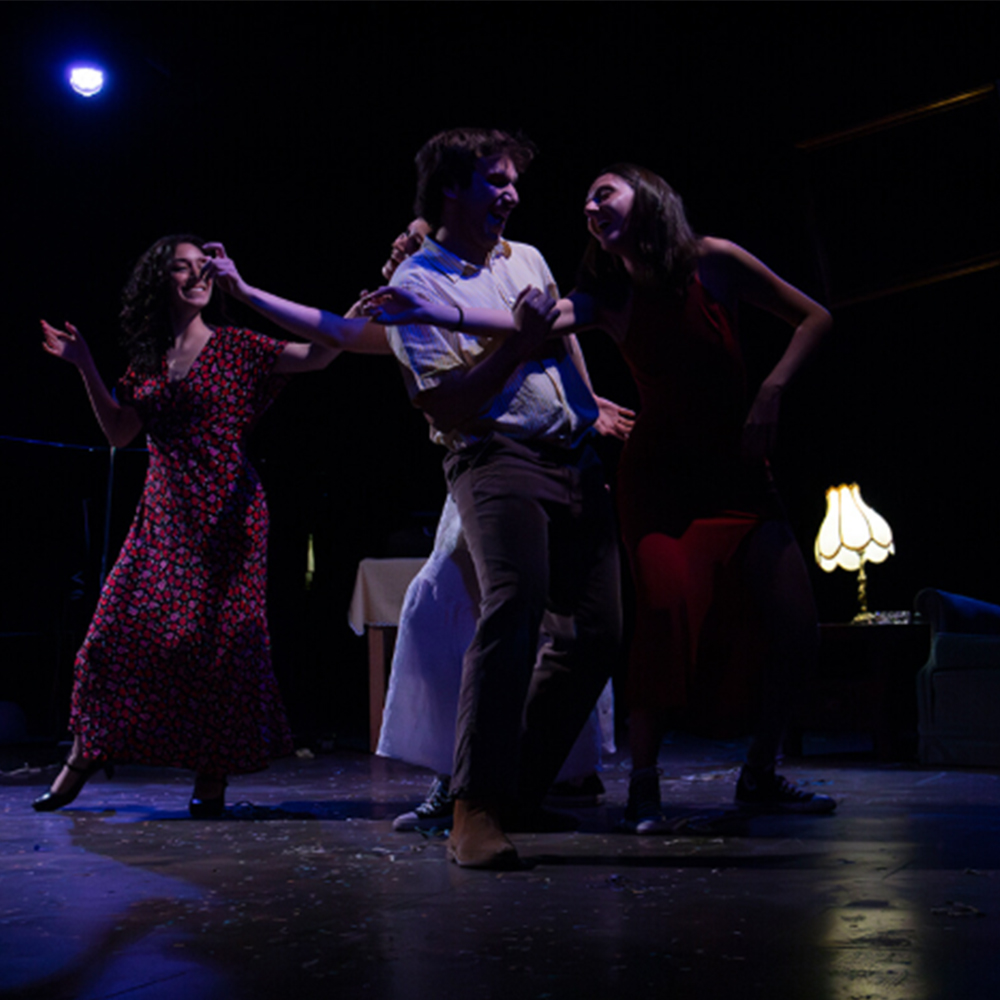 Escena teatral nocturna i dinàmica d'una obra de teatre musical, amb tres personatges en acció al centre. Un home jove amb camisa i pantalons marrons clars està sent agafat o sacsejat per dues dones, una a cada costat. La dona de l'esquerra porta un vestit llarg vermell, té el cabell arrissat fosc i una expressió exuberant i amb un somriure molt obert. La dona de la dreta, amb un vestit vermell fosc i sabatilles esportives fosques, riu mirant cap amunt.