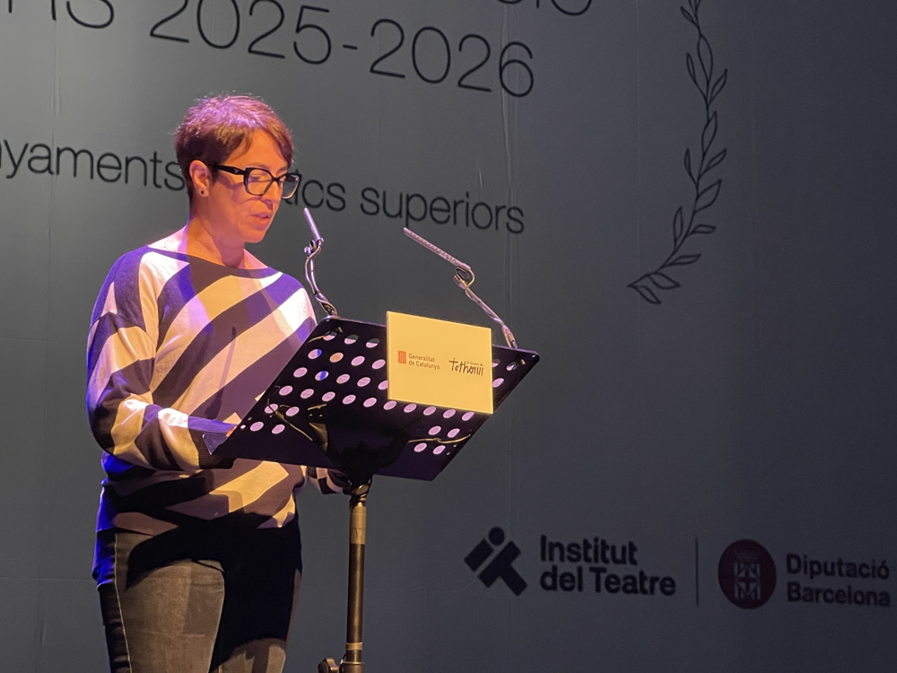 Directora General de l’Institut del Teatre: Núria Plana Núria Plana amb cabells foscos i curts, ulleres i una samarreta de ratlles horitzontals blanques i negres, està parlant en un escenari fosc. Es troba darrere d'un faristol amb micròfon i fulls, amb els peus de la base visibles, durant l'Acte d'Inauguració del Curs 2025-2026 dels Ensenyaments Artístics Superiors.