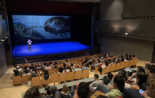 Vista panoràmica d'una sala de teatre o auditori plena de gent assistint a un acte acadèmic. Des d'un punt de vista elevat i lateral, es veu el públic assegut en cadires de fusta i taronja, mirant cap a l'escenari.