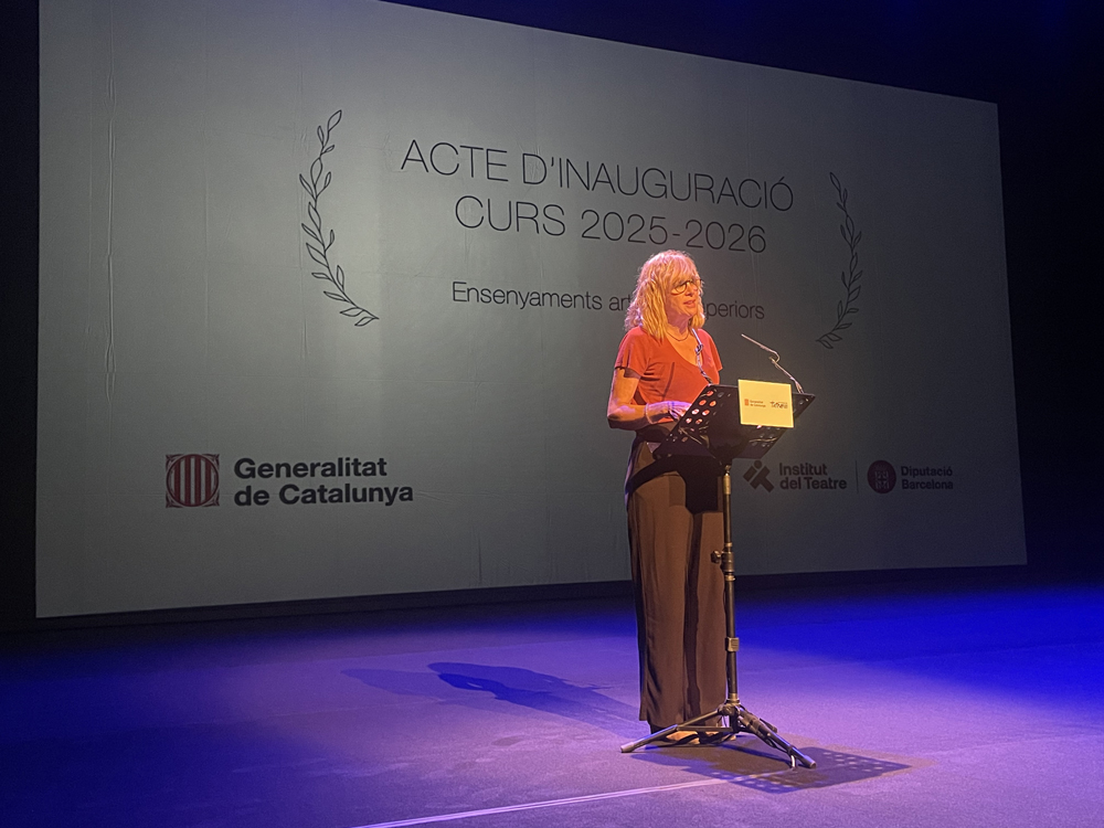 Directora General d’Alumnat: Gemma Verdés Gemma Verdés, amb cabells rossos i un vestit color teula i pantalons foscos, està de peu en un escenari fosc. Està parlant en un faristol amb un micròfon, durant l'acte d'inauguració del Curs 2025-2026 dels Ensenyaments Artístics Superiors. A la part inferior esquerra de la pantalla apareix el logotip de la Generalitat de Catalunya i, a la part inferior dreta, els logotips de l'Institut del Teatre i la Diputació de Barcelona.