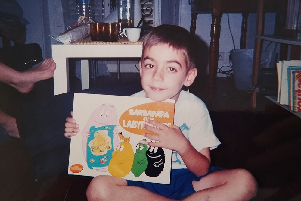 Foto retro, presa en interior, d'un nen petit assegut amb les cames creuades a terra. El nen, amb el cabell fosc i samarreta blanca i pantalons curts blaus, mira directament a càmera amb una lleu somriure. Sosté amb les dues mans un llibre infantil de la col·lecció 'Barbapapa i els Laberints', on es veuen els personatges.