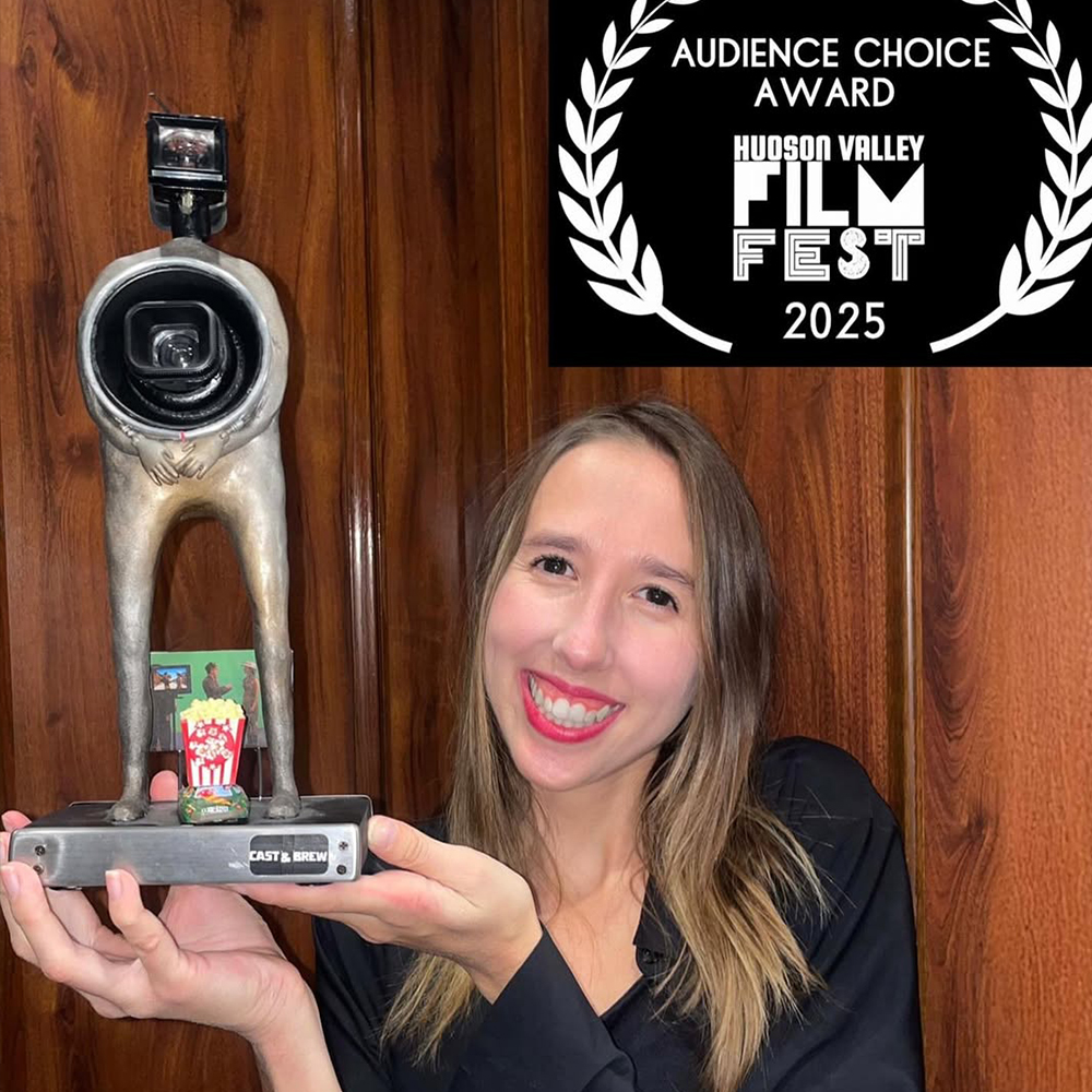 Actriu Claud Hernandez sostenint el guardó Audience Choice Award (Premi del Públic) guanyat al Hudson Valley Film Festival 2025. El trofeu mostra una figura amb un objectiu de càmera com a cap i una base amb el logotip del festival i la marca Cast & Brew.