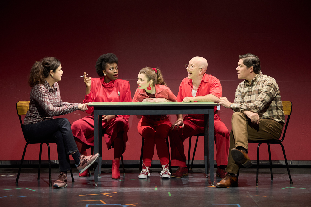 Escena de l'obra de teatre "Com vaig aprendre a conduir", on cinc actors estan asseguts al voltant d'una taula sobre un escenari amb fons vermell.