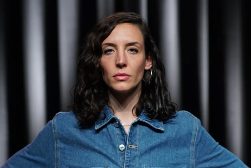 Retrat frontal de l'actriu Berta Pipó mirant fixament a la càmera amb una expressió seriosa i decidida. Vesteix una jaqueta texana de color blau cordada fins al coll i porta els cabells foscos i ondulats per sobre de les espatlles, amb una arracada de cèrcol visible.