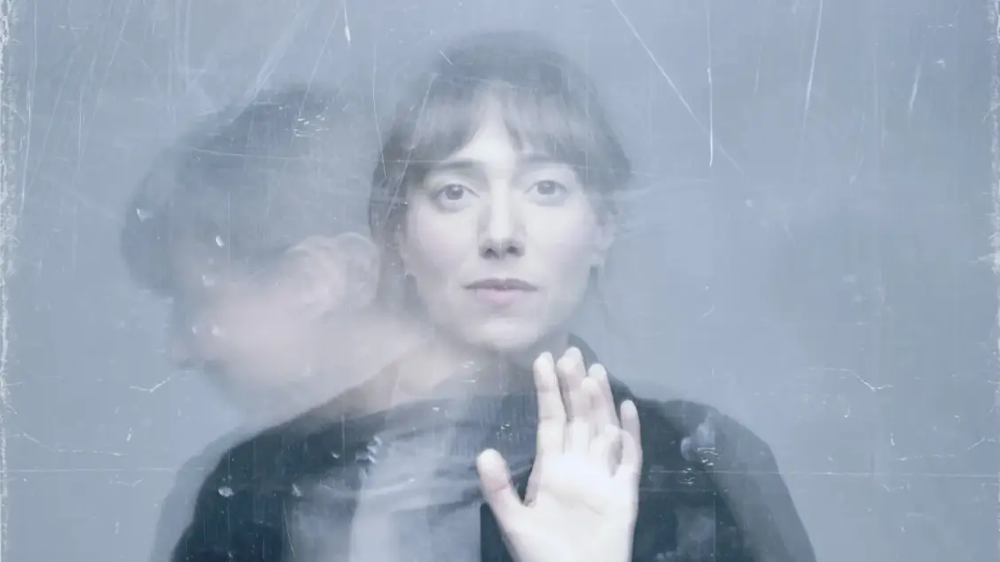 Retrat artístic d'una dona jove, l'Anna Maruny, vist a través d'una superfície translúcida i ratllada que crea un efecte de vidre o plàstic desgastat. La imatge presenta una doble exposició o reflex on es veu el seu rostre de front amb una mirada serena i, simultàniament, el seu perfil desdibuixat.