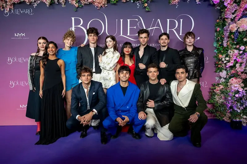 Un gran grup de joves actors i actrius de la sèrie "Boulevard" posen somrients en un photocall esponsoritzat per NYX Professional Makeup. L'escenari té un fons morat fosc amb el títol de la sèrie escrit en lletres grans i blanques al centre i una columna de flors de color rosa i lila a la dreta. Els assistents porten una gran varietat de vestits de gala elegants i de moda, des de vestits llargs negres i vermells fins a jaquetes de color blau vibrant.