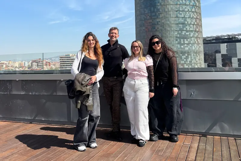 Disseny Hub EÒLIA CSAD 2026 Fotografia de grup en una terrassa assolellada amb vistes a la ciutat de Barcelona. Quatre persones posen davant d'una barana de vidre: una dona amb jaqueta blanca i texans grisos, un home amb jersei negre, una dona amb un polo rosa i pantalons blancs, i una dona amb jersei de xarxa negre i ulleres de sol.