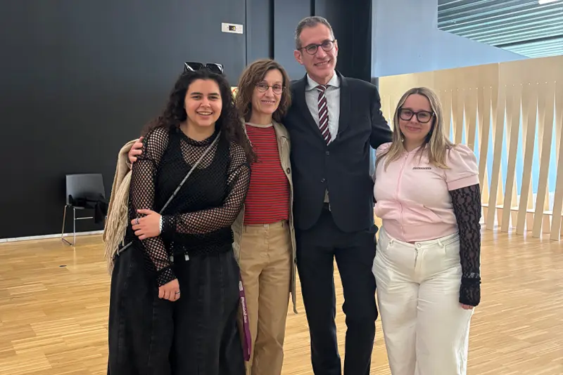 Disseny-Hub-EÒLIA-CSAD Fotografia de grup en un espai interior modern amb terra de fusta. Quatre persones posen somrients davant la càmera. D'esquerra a dreta: una dona jove amb jersei de xarxa negre i faldilla llarga; una dona adulta amb ulleres, jersei de ratlles i jaqueta beix; un home amb vestit fosc, camisa i corbata de ratlles; i una dona jove amb samarreta rosa estil polo i pantalons blancs.