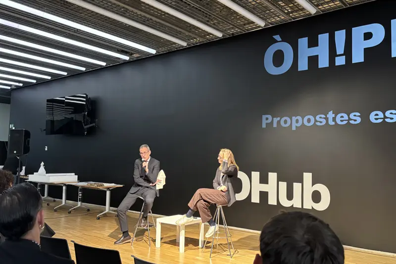 Hub-EÒLIA-CSAD Una conferència o presentació al Disseny Hub Barcelona. Dues persones, un home amb vestit fosc i una dona amb americana i pantalons marrons, estan assegudes en tamborets alts en un escenari amb terra de fusta. L'home parla per un micròfon mentre la dona l'escolta. Al fons, una gran paret negra mostra el text blanc "ÒH!PERA" i "Propostes escèniques".