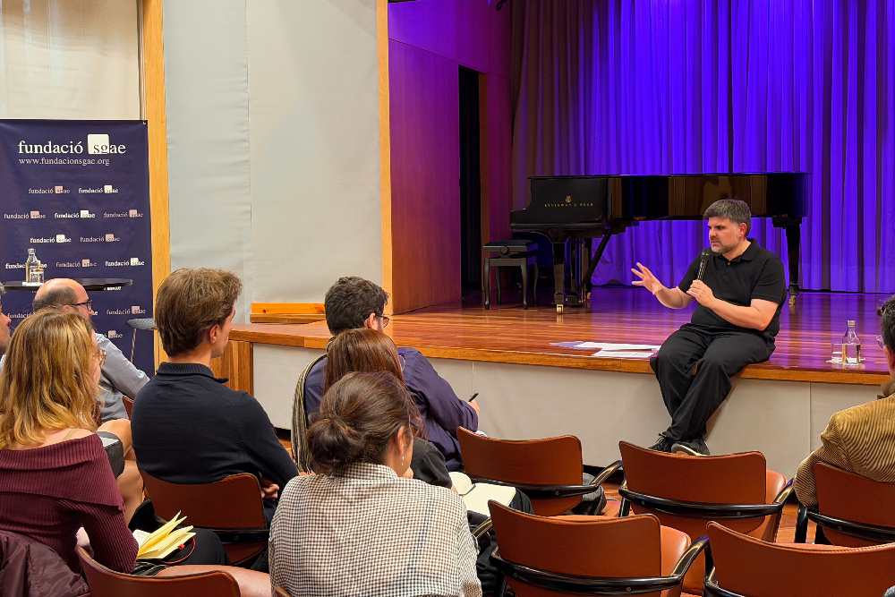 Jordi Casanovas-SGAE Jordi Casanovas assegut a la vora d'un escenari amb un micròfon a la mà, adreçant-se a un públic atent durant un acte de la Fundació SGAE, amb un piano de cua i cortines liles al fons.