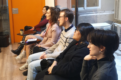 Masterclass Cristina Baggio Un grup de cinc joves asseguts en fila durant una sessió de formació o conferència en una sala amb terra de fusta. Els assistents miren atentament cap a la mateixa direcció, amb actitud d'escolta activa.