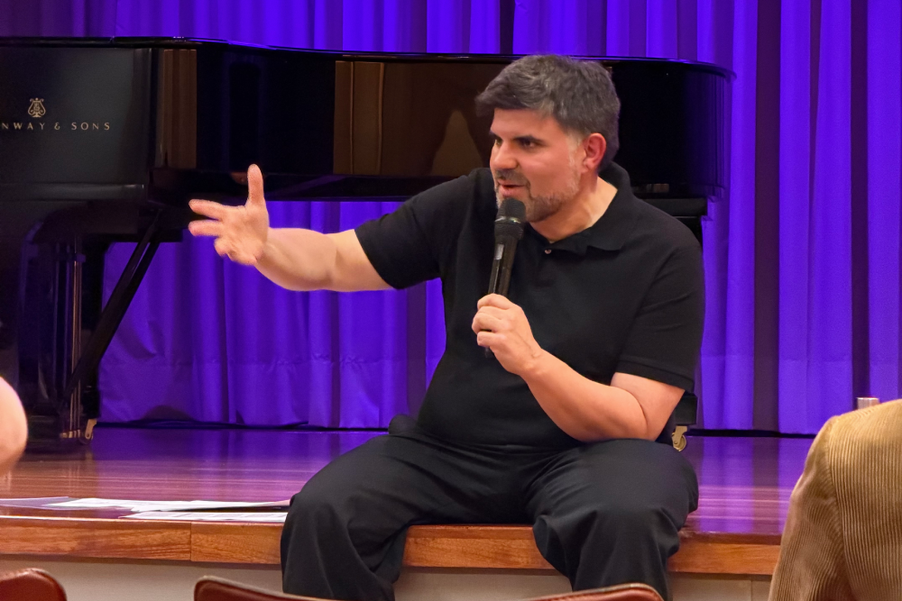 Masterclass Jordi Casanovas Jordi Casanovas assegut a l'escenari mentre parla per un micròfon i gesticula amb la mà durant una xerrada, amb un piano de cua i teló lila al fons.