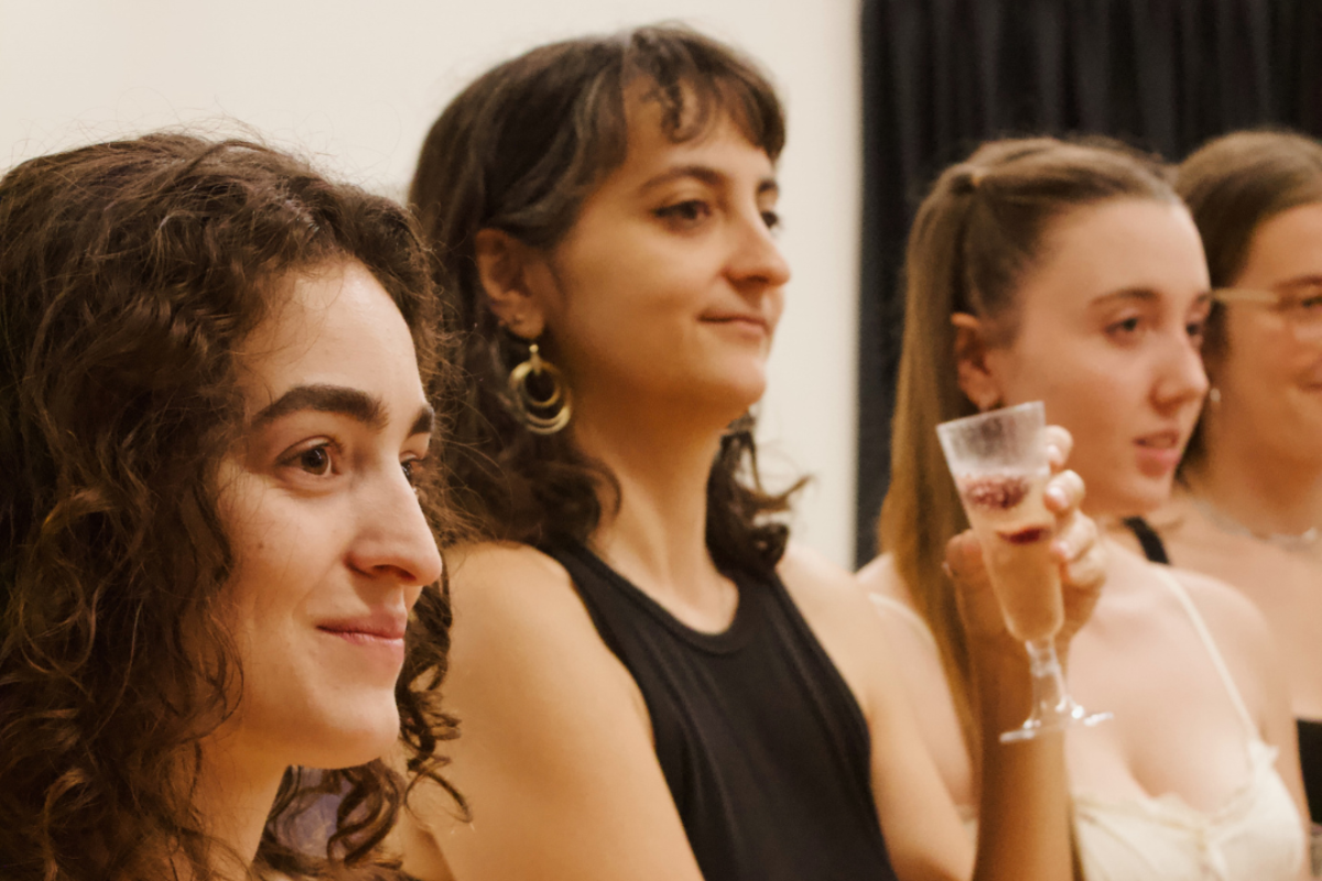 Primer pla de quatre dones joves somrients, de peu i mirant cap a un costat en un acte social. La dona en primer terme, amb cabell arrissat i vestit beix, té els braços creuats. La tercera dona començant per l'esquerra sosté una copa de cava.