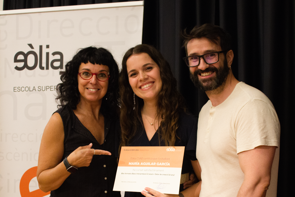 Una estudiant sosté el seu diploma de graduació de la Formació Professional Artística 2026, mentre somriu al costat d'una docent que assenyala el document i un altre membre del personal del centre, davant un bàner de l'Escola Sòlia.