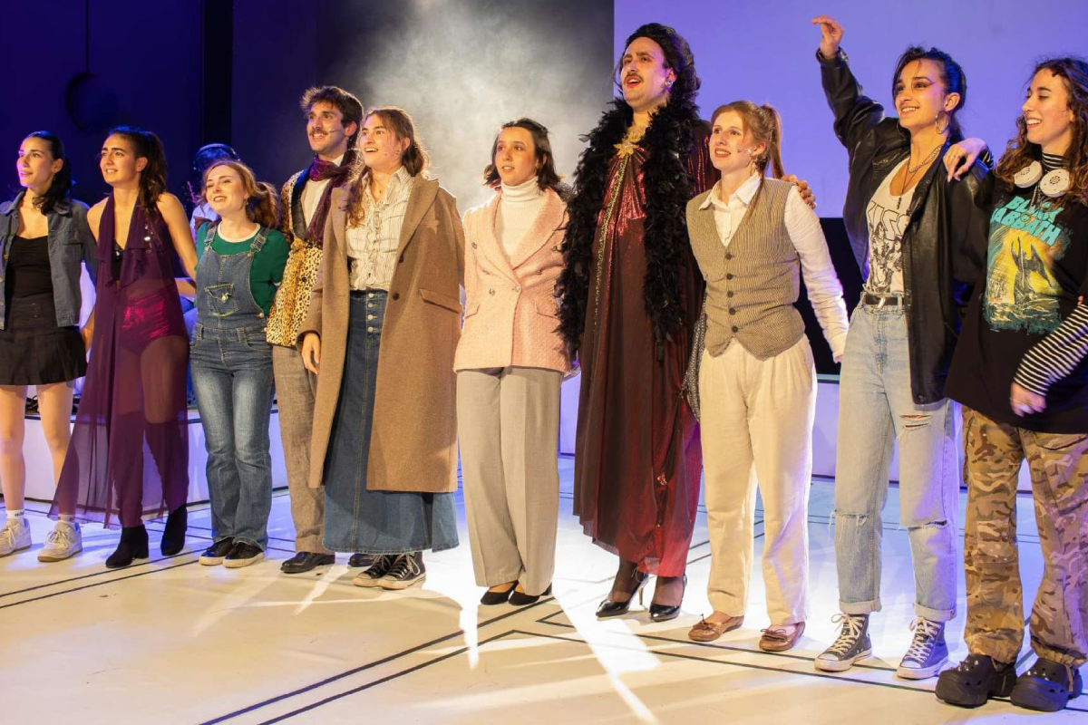 Fotografia de grup dels intèrprets del musical "No es pot saber" saludant somrients sobre l'escenari del Teatre Eòlia, lluint el vestuari divers de la funció.
